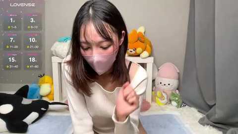 Media: A video shows an Asian woman with shoulder-length black hair and a pink mask, kneeling on a white shag rug, holding a pen. The room is decorated with plush toys, a calendar, and gray curtains.