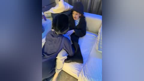 Media: Video of a young boy in a black hoodie, sitting on a beige couch, smiling at a young girl with short black hair, both indoors.