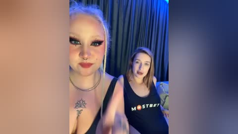 Media: Video of two women in a dimly lit room. Blonde woman with tattoos and piercings, wearing black top, stands beside brunette in black \"MOTHER\" tank top.