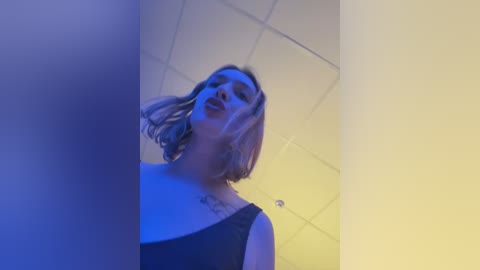 Media: Video of a blonde woman with shoulder-length hair, wearing a black top, standing in a dimly lit room with blue and yellow lighting, ceiling tiles visible.