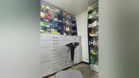 Media: Video of a well-lit, organized pharmaceutical storage room with white cabinets, shelves packed with colorful medication boxes, and a black stool in the center.