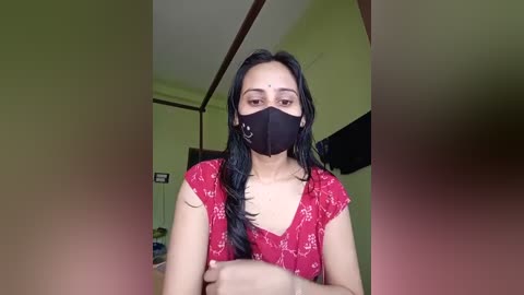 Media: Video of a South Asian woman with long black hair, wearing a red floral dress, black mask, and gold earrings, standing indoors with a green wall and a TV.