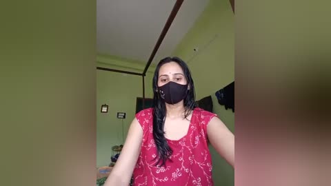 Media: A video of a woman with long black hair, wearing a red T-shirt, black mask, and green walls. Background shows a bed, TV, and framed photos.