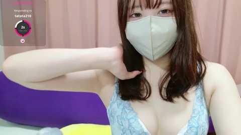 Media: A video of a young Asian woman with long brown hair and a light complexion, wearing a light blue lace bra and a light gray face mask, adjusting her hair while seated on a bed with purple and yellow pillows.