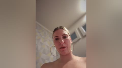 Media: A candid video of a topless woman with blonde hair, wearing large hoop earrings, standing in a bathroom with a white, patterned wallpaper and a window above.