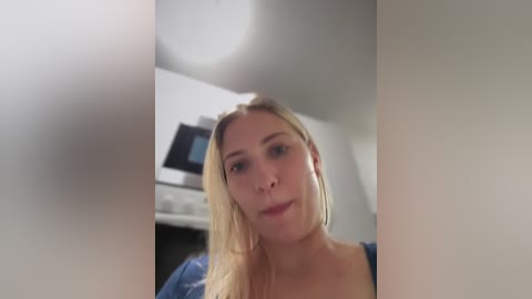 Media: Video of a blonde woman with fair skin and medium build, wearing a blue top, standing in a modern kitchen with white cabinets and a microwave.