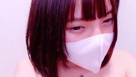 Media: Video of an East Asian woman with straight black hair and bangs, wearing a white face mask and a light-colored top, against a plain white background.