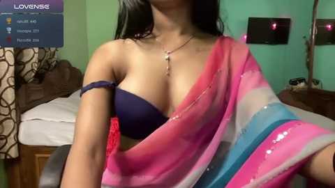 Media: A video of an Indian woman with medium skin tone, long black hair, wearing a dark blue bra and a pink and white saree, sitting in a green room with a bed, curtains, and a TV.