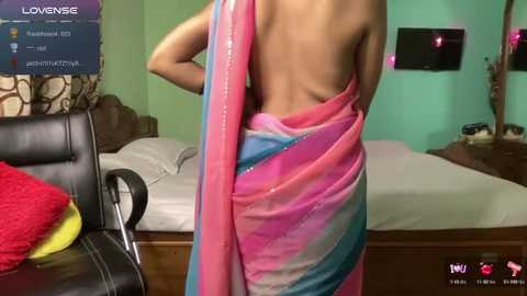 Media: Video of a woman in a colorful, sheer saree with pink, blue, and green stripes, standing in a bedroom with a bed, green walls, and a flat-screen TV.