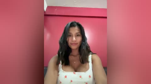 Media: Video of a Latina woman with long black hair, wearing a white tank top with red strawberries, standing in a red-walled room with a ceiling fan.