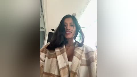 Media: Video of a young woman with long, dark hair, wearing a beige and white plaid bathrobe, standing in a bright, minimalist room with white walls and a window.