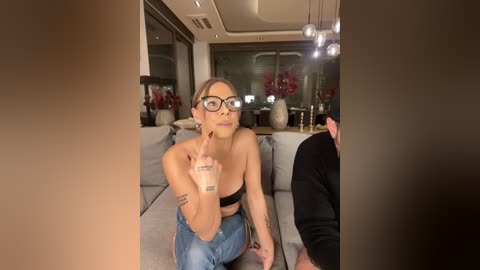 Media: A video of a blonde woman with glasses, tattoos, and a black bra, sitting on a sofa, flipping the bird, in a modern, dimly-lit living room with large windows and decorative vases.