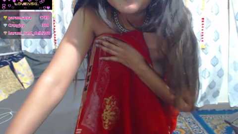 Media: A video of a young woman with long, dark hair partially wrapped in a red saree, holding it up with her right hand. She is in a room with white curtains and a blue and yellow rug.