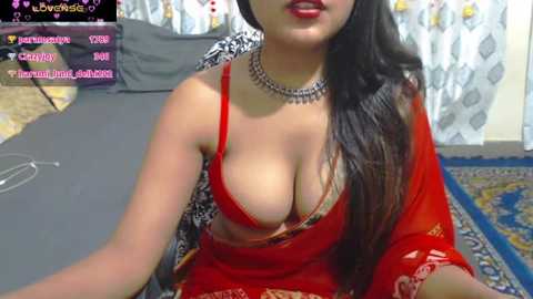 Media: A video of a South Asian woman with long dark hair, wearing a red saree with a low-cut blouse revealing ample cleavage, sitting on a bed. The background features a patterned wall and a TV screen with streaming details.