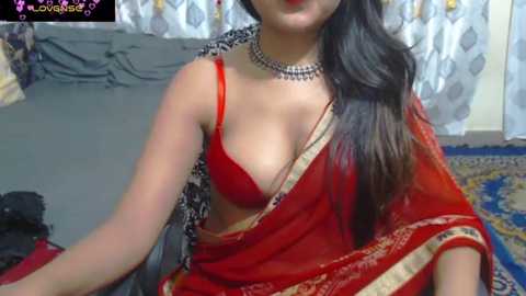 Media: A video of a South Asian woman in a red saree with a gold border, revealing a red bra, sitting indoors on a patterned rug. She wears a black choker necklace and has long, dark hair.