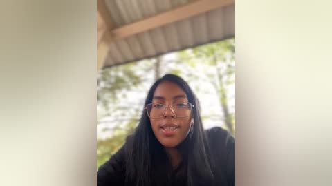 Media: A video of a young Black woman with long, straight black hair, wearing glasses, and a black sweater, standing in a rustic wooden structure with a greenery backdrop.