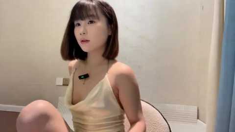 Media: A video of an East Asian woman with short brown hair and fair skin, wearing a beige, low-cut, sleeveless top, sitting on a bed in a simple room with beige walls.