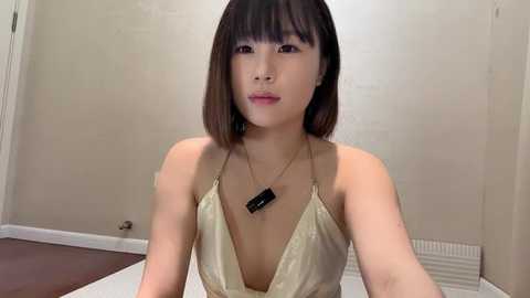 Media: Video of an Asian woman with straight, shoulder-length black hair and bangs, wearing a gold halterneck dress, standing indoors against a beige wall, with a radiator visible below.