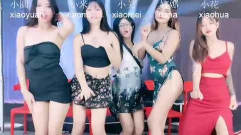 Media: Video of five Asian women in casual outfits, including black strapless, floral shorts, and a red dress, posing with joyful expressions against a modern indoor setting with red chairs.