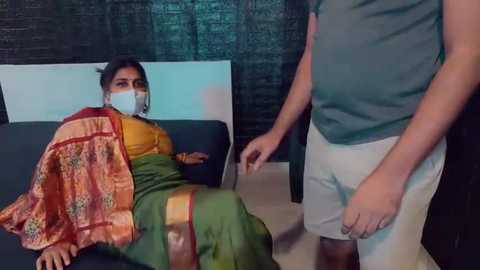 Media: Video of a woman in a yellow blouse and green sari lying on a bed, wearing a mask, while a man in a gray shirt and white pants stands beside her.