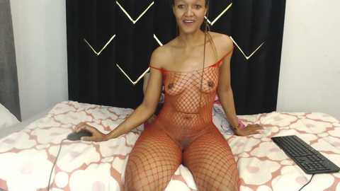 Media: Video of a nude, light-skinned woman with medium-length braided hair, wearing a red fishnet bodysuit, seated on a bed with a black curtain backdrop.