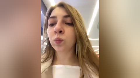 Media: Video of a young woman with long, light brown hair, wearing a white top, making a kissy face. She has fair skin and is indoors with a white ceiling and blurred background.