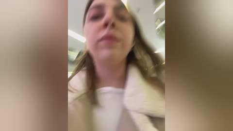 Media: Video of a woman with fair skin, long brown hair, wearing a white shirt, slightly blurry, taken from a low angle, indoors with fluorescent lighting.