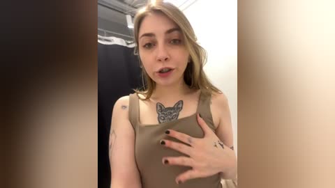 Media: Video of a young Caucasian woman with shoulder-length blonde hair, wearing a sleeveless beige top, and a prominent tattoo of a wolf on her chest. She has a neutral facial expression and black-painted nails. The background shows a shower stall with a white curtain and a beige wall.