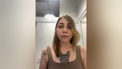 Media: Video of a young woman with fair skin and light brown hair, wearing a brown tank top, standing in a minimalist, white-walled bathroom with a large wolf tattoo on her chest.