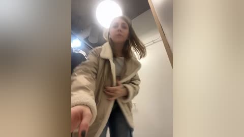 Media: Video of a young woman with straight, shoulder-length light brown hair, wearing a beige fur-lined coat and blue jeans, reaching out to open a door. Background features a white wall and a ceiling light.