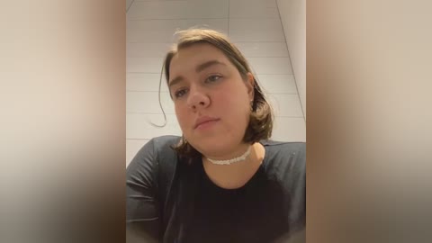 Media: Video of a young Caucasian woman with short, light brown hair and a light complexion, wearing a black top and white choker necklace, standing in a white-tiled hallway.