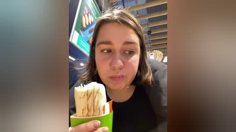 Media: Video of a young woman with light brown hair, wearing a black top, eating ice cream in a green cup, indoors with blurred people in the background.