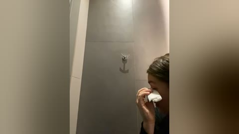 Media: Video of a person with light skin and brown hair, wearing a black shirt, blowing their nose in a public restroom with gray tiled walls and a metal hook.