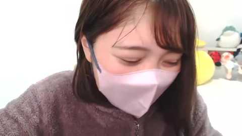 Media: Video of an Asian woman with straight brown hair, wearing a gray fleece jacket and a white mask, eyes closed, possibly resting. Background features stuffed toys and a white wall.