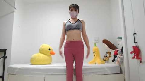 Media: Video of a young Asian woman in a white mask, gray sports bra, and pink leggings, standing in a minimalist, white room with a bed, yellow duck toys, and stuffed animals.