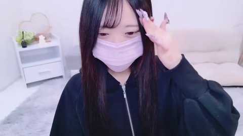 Media: A video of an Asian woman with long black hair, wearing a pink face mask, black jacket, and black gloves, adjusting her hair in a minimalist, modern living room with white furniture and a grey rug.