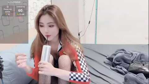 Media: A video of a young East Asian woman with straight brown hair, wearing a colorful patterned jacket, fishnet stockings, and drinking from a silver straw. She sits on a bed with messy clothes in a simple, brightly-lit room.