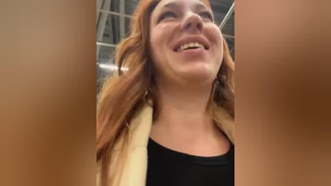 Media: Video of a smiling woman with long, wavy red hair, wearing a black top, taken from a low angle, showing her face and upper body in a beige sweater.