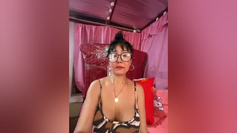 Media: Video of a young Asian woman with black hair in a high bun, wearing glasses and a leopard-print bikini top, sitting in a pink tent with red bedding, large hoop earrings, and a red pillow.