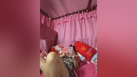 Media: Video of a young woman lying on a bed with pink curtains, wearing a leopard-print top, holding a smartphone, surrounded by stuffed toys and colorful pillows.