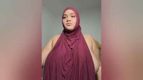 Media: Video of a plus-size woman with light brown skin, wearing a maroon hijab, sitting in a neutral indoor setting.