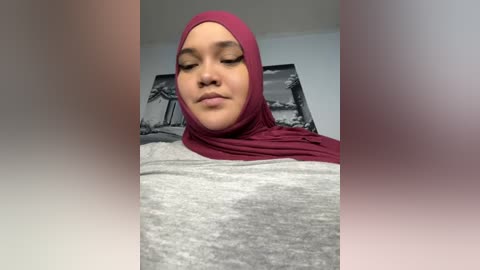 Media: Video of a woman with light brown skin and a maroon hijab, wearing a gray and white top, indoors with a black-and-white palm tree wall art in the background.