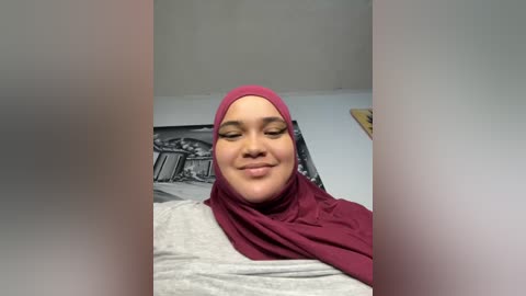 Media: Video of a smiling woman with light brown skin, wearing a maroon hijab and light gray top, in a dimly lit room with a black and white abstract wall hanging.