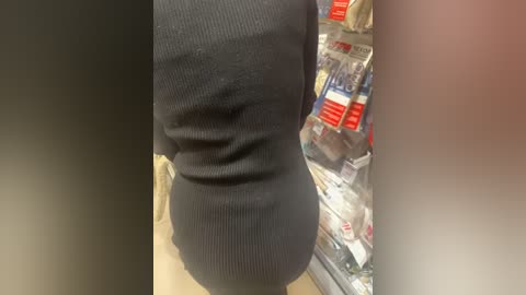 Media: Video of a person from the back, wearing a black ribbed sweater, standing in a grocery store aisle filled with red-labeled packaged food items and plastic bags.