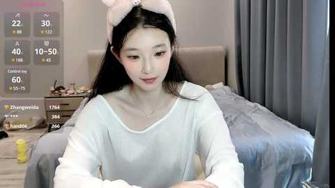 Media: Video of an East Asian woman with long black hair and fair skin, wearing a white hoodie, sitting in a modern bedroom with a bed, nightstand, and digital temperature display.