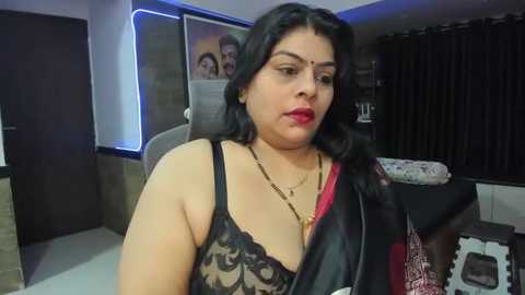 Media: Video of a middle-aged South Asian woman with medium skin tone, wearing a black lace bra, red lipstick, and traditional jewelry. She sits in a modern living room with dark curtains and a large TV in the background.