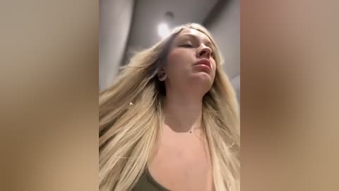 Media: Video of a blonde woman with long, wavy hair, wearing a green tank top, standing in a modern, minimalist room with beige walls and recessed lighting. Her eyes are closed, and she appears relaxed.