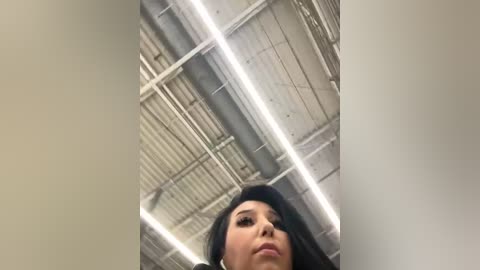 Media: Video of a young woman with long black hair and a serious expression, captured from below, in an industrial setting with metallic ceilings and exposed pipes.