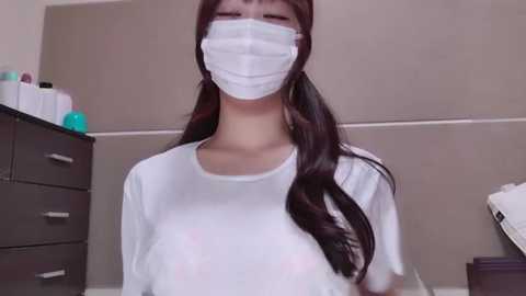 Media: Video of an East Asian woman with long black hair, wearing a white mask and t-shirt, standing in a bathroom with beige walls, dark drawers, and toiletries.