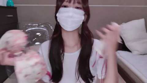 Media: A video of an Asian woman with long black hair, wearing a white surgical mask, standing in a hospital room. She is holding a bloody cloth.